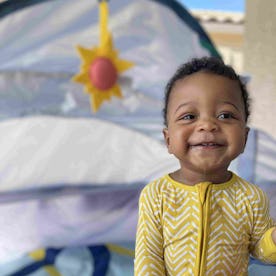 A joyful toddler in yellow pajamas holding a "My 1st ABC" book in front of a play tent with a sun de...